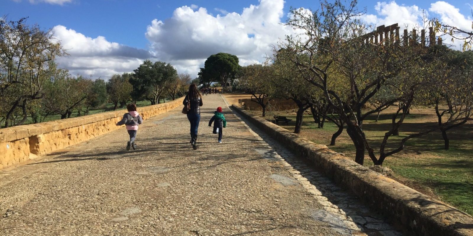 Mamma e due bambini che corrono lungo la via sacra tra mandorli e ulivi verso il tempio di Giunone sullo sfondo sotto un cielo azzurro con nuvole bianche