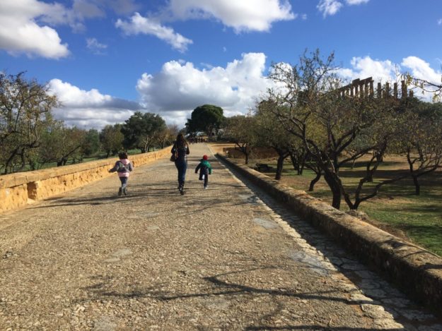 Mamma e due bambini che corrono lungo la via sacra tra mandorli e ulivi verso il tempio di Giunone sullo sfondo sotto un cielo azzurro con nuvole bianche