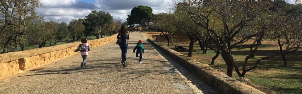 Mamma e due bambini che corrono lungo la via sacra tra mandorli e ulivi verso il tempio di Giunone sullo sfondo sotto un cielo azzurro con nuvole bianche